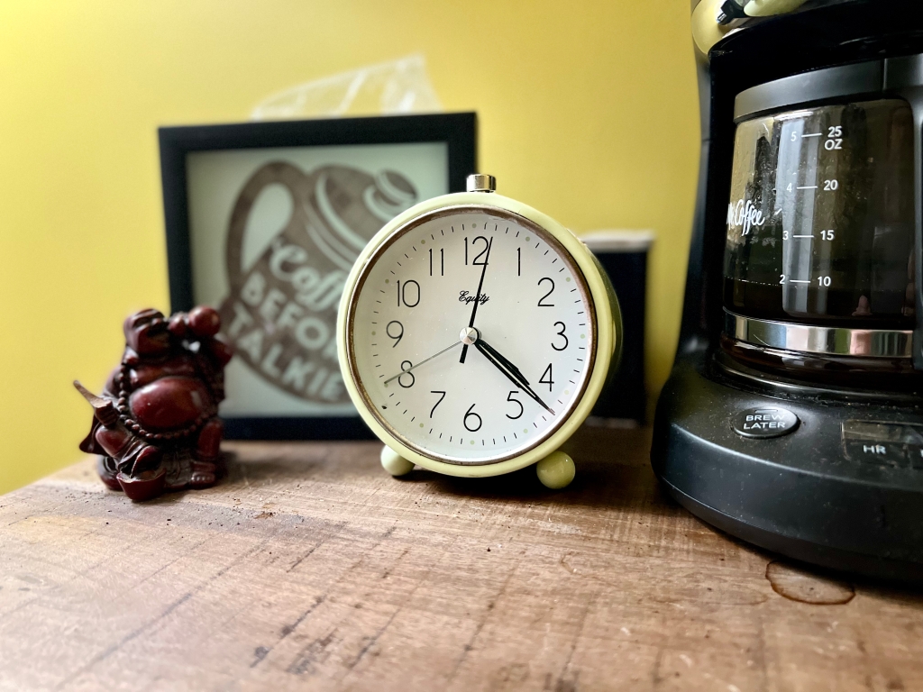 A picture of an analog alarm clock next to a coffee maker, a Buddha, and a sign from Target that says "Coffee Before Talkie."